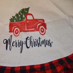 May include: A Christmas tree skirt with a red truck carrying a green Christmas tree. The words "Merry Christmas" are written in a script font. The skirt has a red and black buffalo plaid border.