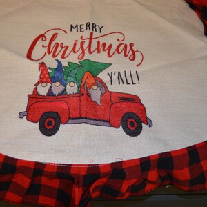 May include: A Christmas-themed tree skirt with a red and black buffalo plaid ruffle. The center features a red truck carrying gnomes and a Christmas tree, with the text "MERRY Christmas Y'ALL!" in red script.