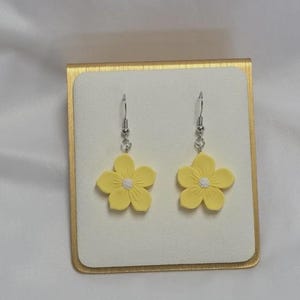 May include: A pair of yellow flower earrings with white centers and silver hooks. The earrings are displayed on a white surface inside a gold-colored box.