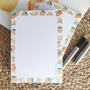 Pode incluir: Papelaria branca com um design em aquarela de pêssegos e limões. A borda do papel é decorada com pêssegos e folhas verdes. Duas canetas estão em uma superfície tecida. O texto "La Boutique à Souvenirs" é visível.