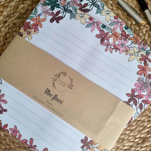Peut inclure: Un bloc-notes fleuri avec des pages lignées et une bordure décorative de fleurs colorées. Le bloc-notes est enveloppé dans du papier brun et porte l'inscription "Bloc fleuri". Le bloc-notes est posé sur une surface tissée.