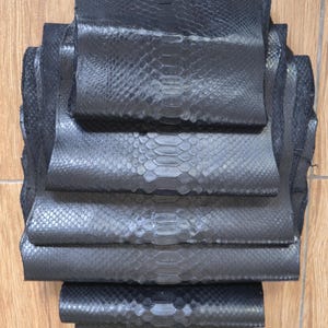 Op de afbeelding: Een stapel zwarte slangenleren stukken met een duidelijk, herhalend schubbenpatroon. Het materiaal heeft een matte afwerking en is in een verticale opstelling gelaagd, wat de natuurlijke textuur en de langwerpige vorm van de huid laat zien.