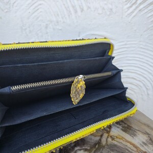 Puede incluir: Una cartera de piel de serpiente amarilla con cierre de cremallera y un interior negro. La cartera est&aacute; abierta y el tirador de la cremallera es visible.