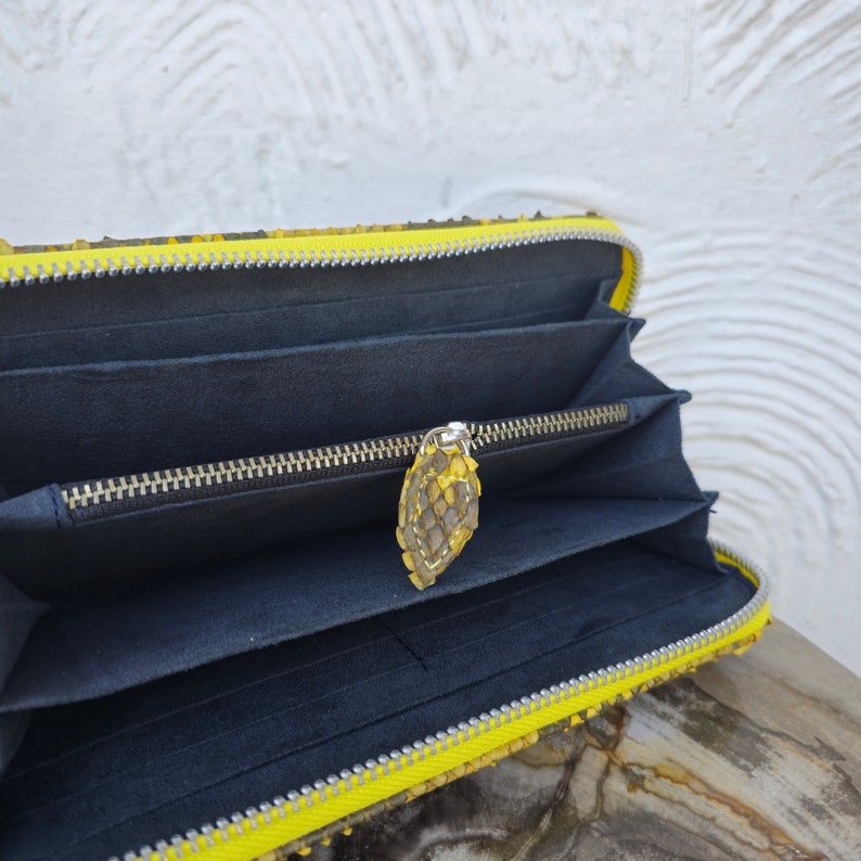 Puede incluir: Una cartera de piel de serpiente amarilla con cierre de cremallera y un interior negro. La cartera est&aacute; abierta y el tirador de la cremallera es visible.
