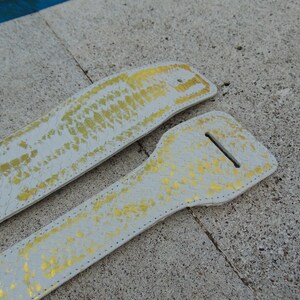 Könnte beinhalten: Zwei weiße Ledergitarrenriemen mit einem goldenen Schlangenhautmuster. Die Riemen liegen flach auf einer hellgrauen Oberfläche.