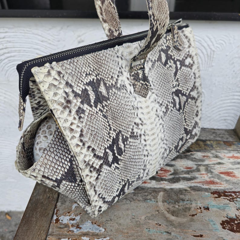 Peut inclure: Un sac &agrave; main en cuir de serpent blanc et gris avec des accents noirs et une poign&eacute;e sup&eacute;rieure.