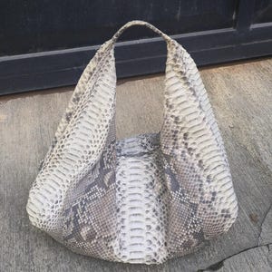 May include: A hobo-style handbag crafted from snakeskin featuring a natural scale pattern in shades of cream, grey, and charcoal. The bag has a single integrated handle and a slouchy, unstructured silhouette with a textured, overlapping scale finish.
