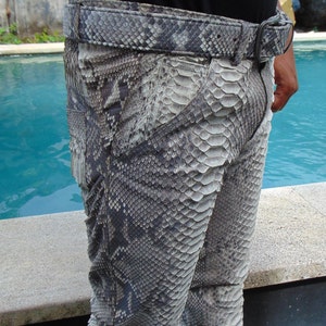 K&ouml;nnte beinhalten: Grau-schwarz gemusterte Hose und G&uuml;rtel. Die Hose und der G&uuml;rtel haben ein Schlangenhaut-Design. Der G&uuml;rtel hat eine silberne Schnalle. Die Hose wird mit einem schwarzen Oberteil getragen. Die Hose besteht aus einem Material, das Schlangenhaut &auml;hnelt.