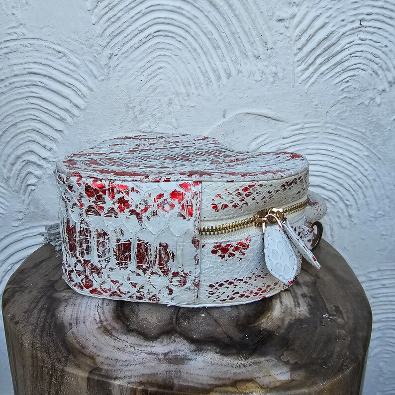 Pu&ograve; includere: Un beauty case a forma di cuore con un motivo a pelle di serpente bianco e rosso e una cerniera dorata. La borsa &egrave; appoggiata su una superficie di legno. Dimensioni: circa 15 cm di diametro.