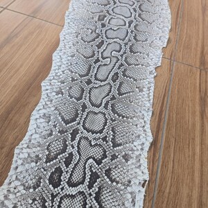 May include: A long, shed snake skin featuring a repeating pattern of dark grey and black blotches against a white, textured background. The scales are clearly defined, creating a natural, organic texture that runs the length of the specimen.