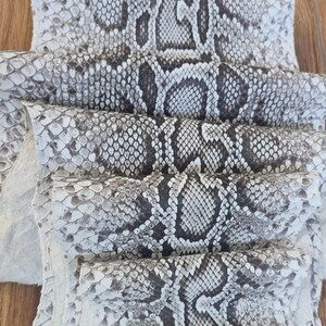 May include: A stack of multiple pieces of snakeskin leather featuring a repeating scale pattern in shades of white, grey, and dark brown. Each piece of hide is layered to display the natural texture and the distinct, organic markings of the reptile skin.