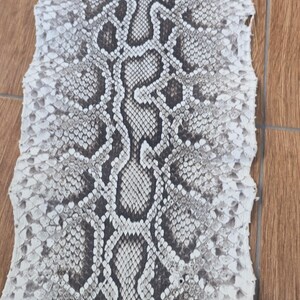 May include: A flat, elongated piece of snakeskin featuring a repeating pattern of dark grey and black diamond-shaped scales centred against a lighter, off-white background. The edges of the skin are irregular and textured with small, overlapping scales.