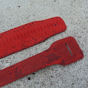 Könnte beinhalten: Zwei rote Gitarrengurte aus Leder mit Schlangenhautmuster. Die Gurte liegen flach auf einer grauen Betonfläche.