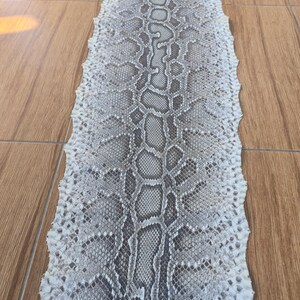 May include: A long, flat, tanned snake skin featuring a repeating diamond-shaped pattern in shades of off-white, light gray, and dark charcoal. The texture is visibly scaled, with a jagged, uneven edge running along the entire length of the hide.