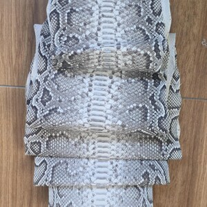 K&ouml;nnte beinhalten: Ein Stapel nat&uuml;rlicher Schlangenhaut-Lederh&auml;ute mit einem sich wiederholenden Rautenmuster in Off-White, Grau und Dunkelbraun. Die texturierten Schuppen sind in Schichten angeordnet und zeigen den markanten R&uuml;ckenstreifen und die organische Struktur.