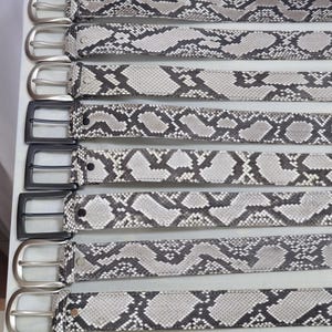 Puede incluir: Una colección de cinturones con estampado de piel de serpiente. Los cinturones presentan un patrón gris y negro con hebillas plateadas y negras. Los cinturones están dispuestos en fila, mostrando los diferentes estilos de hebillas.