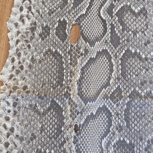 Peut inclure: Une pi&egrave;ce de cuir de peau de serpent rectangulaire et plate pr&eacute;sentant un motif d'&eacute;cailles naturel dans des tons de gris, blanc et anthracite. La peau affiche une texture en losanges r&eacute;p&eacute;titive sur sa surface, avec deux petits trous.