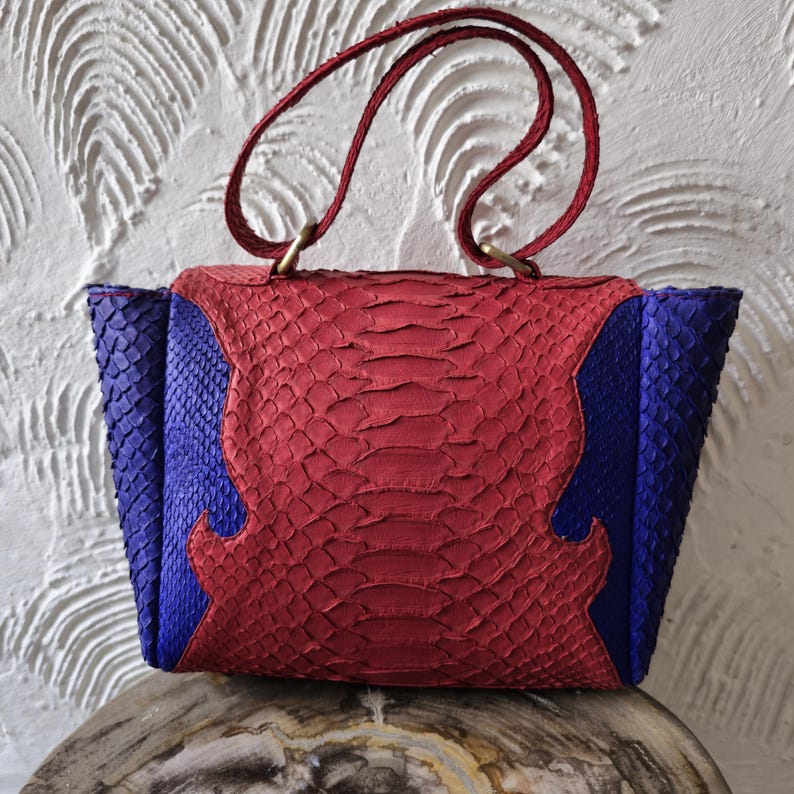 May include: A red and blue snakeskin handbag with two top handles. The bag has a unique design with a contrasting colour pattern.