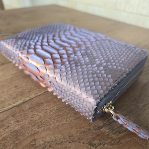 Op de afbeelding: Een lichtpaarse, getextureerde portemonnee met een ritssluiting en een goudkleurige ritstrekker. Het oppervlak van de portemonnee heeft een schubbenpatroon en de ritssluiting is grijs. De portemonnee ligt op een houten oppervlak.