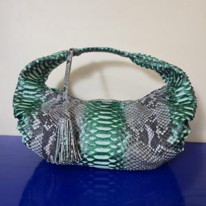 May include: A green, gray, and white snakeskin handbag with a curved handle and a tassel. The bag has a rounded shape and a central panel of vibrant green scales. The handle and tassel are made of the same snakeskin material.