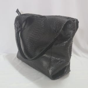 May include: A black tote bag with a snakeskin effect. The bag has a rectangular shape with a large main compartment and two handles. The bag is made of a soft, flexible material.