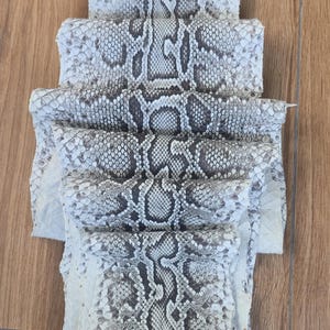 May include: A folded piece of natural snakeskin leather featuring a repeating diamond-shaped scale pattern in shades of off-white, cream, and dark grey. The hide is layered in multiple sections, displaying the intricate texture and organic markings.