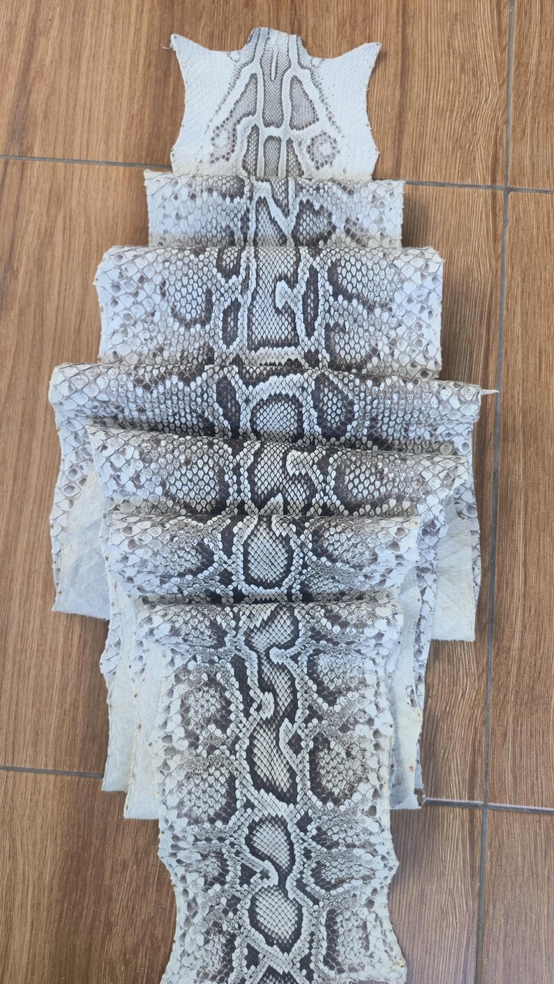 May include: A folded piece of natural snakeskin leather featuring a repeating diamond-shaped scale pattern in shades of off-white, cream, and dark grey. The hide is layered in multiple sections, displaying the intricate texture and organic markings.
