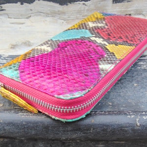 K&ouml;nnte beinhalten: Eine farbenfrohe Schlangenleder-Geldb&ouml;rse mit einem pink-, gelb-, blau- und gr&uuml;nen Muster. Die Geldb&ouml;rse hat einen Rei&szlig;verschluss.