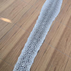 May include: A long, shed snake skin featuring a detailed, repeating scale pattern in shades of white, light grey, and dark grey. The skin is elongated and narrow, displaying a natural, textured surface with a distinct, organic, and intricate design.