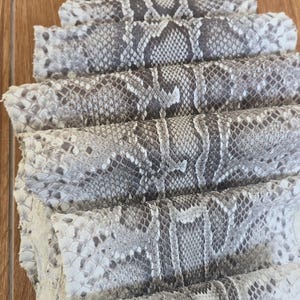 May include: A stack of folded snake skin leather featuring a natural grey, white, and black diamond-shaped scale pattern. The material is layered in multiple horizontal sections, showcasing the detailed texture and organic markings of the reptile hide.