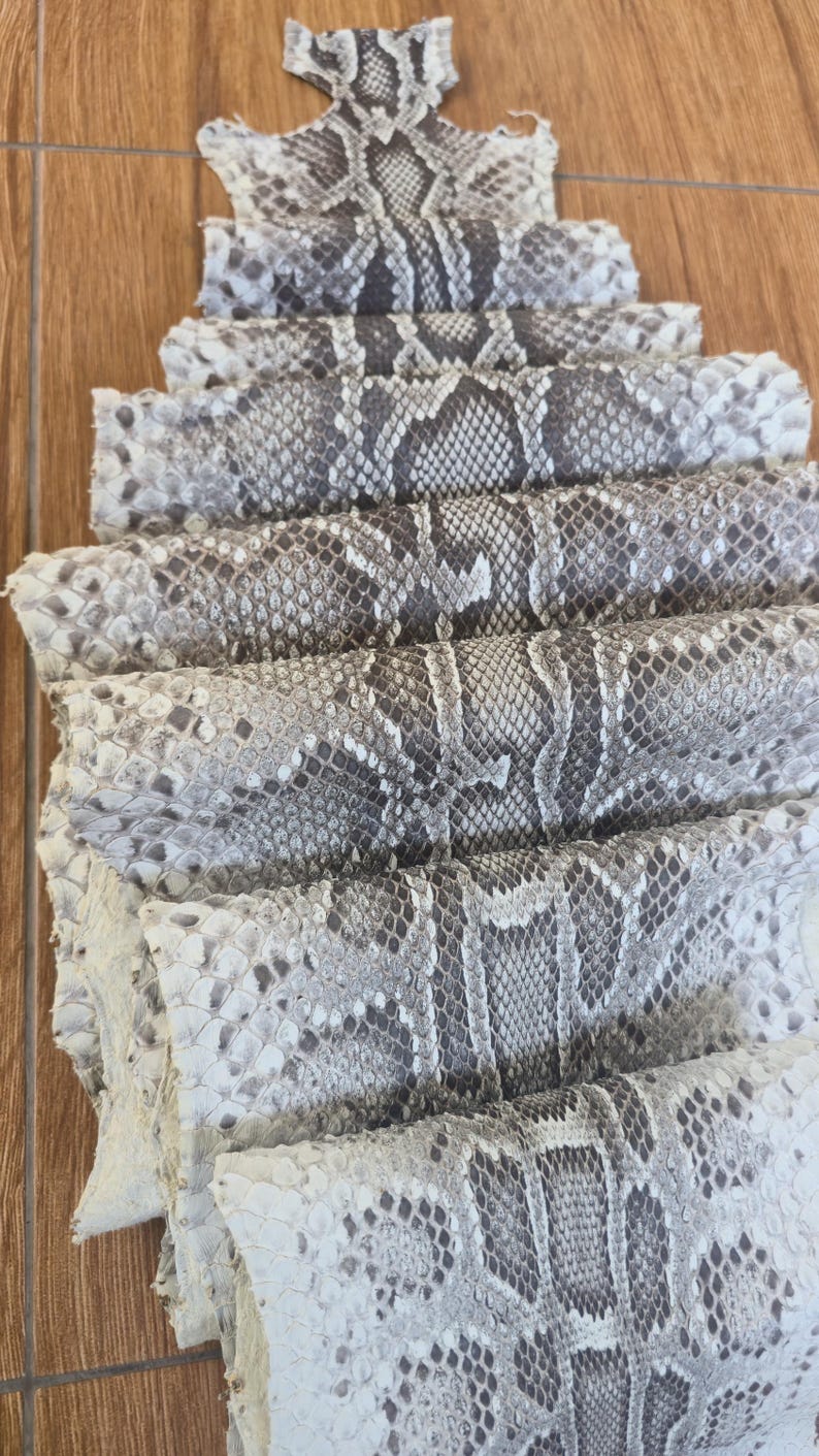 May include: A stack of folded snake skin leather featuring a natural grey, white, and black diamond-shaped scale pattern. The material is layered in multiple horizontal sections, showcasing the detailed texture and organic markings of the reptile hide.