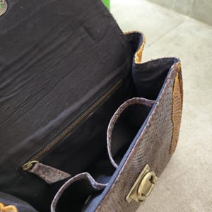 May include: A brown and tan snakeskin handbag with a black fabric interior. The bag has a zipper closure and a brass lock.
