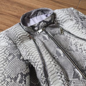 Peut inclure: Une veste avec un motif peau de serpent gris et blanc, dotée d'un col montant, d'une fermeture éclair centrale argentée et de deux poches zippées argentées sur le devant. Le vêtement possède une doublure intérieure de couleur claire.
