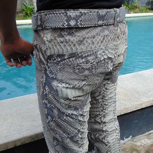 K&ouml;nnte beinhalten: Nahaufnahme einer Hose aus Schlangenhaut mit passendem G&uuml;rtel. Die Hose ist eine Mischung aus Grau, Schwarz und Wei&szlig; mit einem strukturierten, schuppigen Muster. Der G&uuml;rtel hat ein &auml;hnliches Design. Die Hose wird von einer Person getragen.