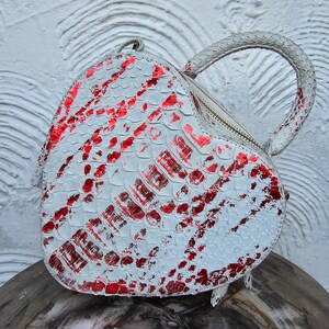 Pu&ograve; includere: Una borsa a forma di cuore con un motivo a pelle di serpente bianco e rosso. La borsa ha una chiusura a cerniera e un manico arrotondato. La borsa &egrave; realizzata in un materiale strutturato.