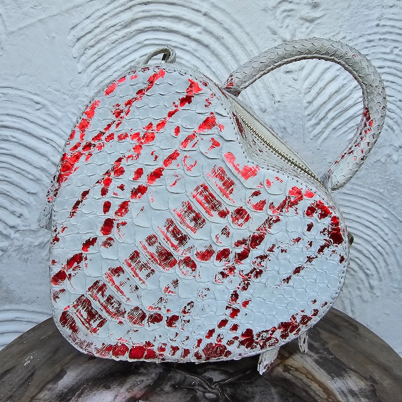 Pu&ograve; includere: Una borsa a forma di cuore con un motivo a pelle di serpente bianco e rosso. La borsa ha una chiusura a cerniera e un manico arrotondato. La borsa &egrave; realizzata in un materiale strutturato.