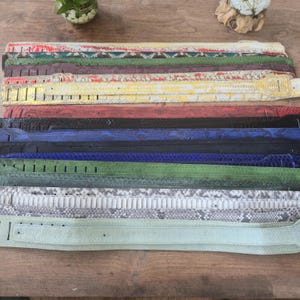 May include: A collection of eleven guitar straps arranged in a stack, featuring various colors including yellow, red, blue, green, black, and grey. Each strap displays a textured snakeskin pattern and includes pre-cut adjustment slots at the ends.