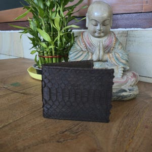 May include: A dark brown leather wallet with a scale-like texture. The wallet is rectangular and appears to be folded. The background includes a statue of a meditating figure and a potted plant.