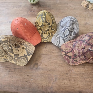 May include: Five baseball caps featuring textured snake skin patterns in various colors including orange, yellow, grey, brown, and a reddish-brown tone. Each cap has a curved brim, a structured crown, and small metal eyelets for ventilation.
