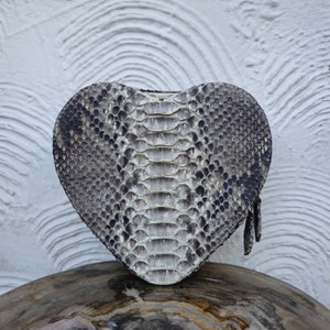 Puede incluir: Bolso de mano de piel de serpiente gris y blanca en forma de corazón con cierre de cremallera. El bolso está hecho de piel de serpiente auténtica y tiene un patrón de escamas único. El bolso de mano está sobre una superficie de madera.