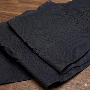 以下が含まれることがあります： 黒い革のクローズアップ。ヘビ革模様。革は折りたたまれ、鱗の質感と詳細が明らかになっています。この素材は柔らかくしなやかで、さまざまな工芸プロジェクトに適しているようです。