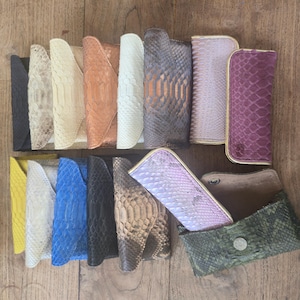 Puede incluir: Una colección de estuches rectangulares para gafas con un patrón texturizado de piel de serpiente en varios colores, incluyendo negro, blanco, beige, azul, amarillo, morado y verde. Cada estuche tiene cierre de solapa, uno está abierto con un botón.