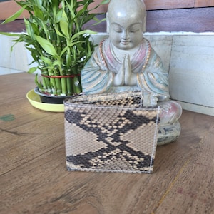 May include: A bi-fold wallet with a snakeskin pattern in shades of tan, black, and gray. The wallet is made of a textured material and is displayed on a wooden surface. A small statue of a meditating figure is in the background.