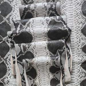 May include: Close-up of several pieces of snakeskin leather. The leather has a pattern of black, gray, and white scales. The pieces are folded and stacked, revealing the texture and color variations of the material. The overall theme is exotic leather goods.