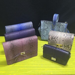 May include: A collection of five handbags and clutches featuring various textures and colors. Three rectangular clutches in blue, purple, and mauve with snakeskin patterns, one blue snakeskin tote, and two smaller structured bags in grey and black.