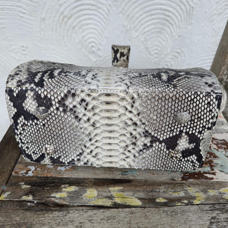 Peut inclure: Un sac &agrave; main en cuir de serpent blanc et noir avec un motif textur&eacute;. Le sac a une poign&eacute;e sup&eacute;rieure et une fermeture &agrave; glissi&egrave;re.