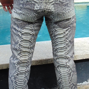 K&ouml;nnte beinhalten: Eine Hose aus grau-wei&szlig;em Schlangenhaut. Die Hose hat einen G&uuml;rtel und Taschen. Das Schlangenhautmuster ist auf der gesamten Hose sichtbar. Die Hose wird von einer Person getragen, die sich in der N&auml;he eines Pools befindet.