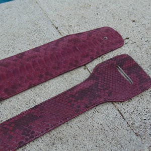 Könnte beinhalten: Zwei burgunderfarbene Gitarrengurte aus Leder mit Schlangenhautmuster. Die Gurte haben eine Metallschnalle und ein Loch zum Einstellen der Länge.