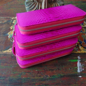 K&ouml;nnte beinhalten: Stapel von vier rosa Ledergeldb&ouml;rsen mit Rei&szlig;verschluss. Die Geldb&ouml;rsen haben ein strukturiertes Schlangenhautmuster.