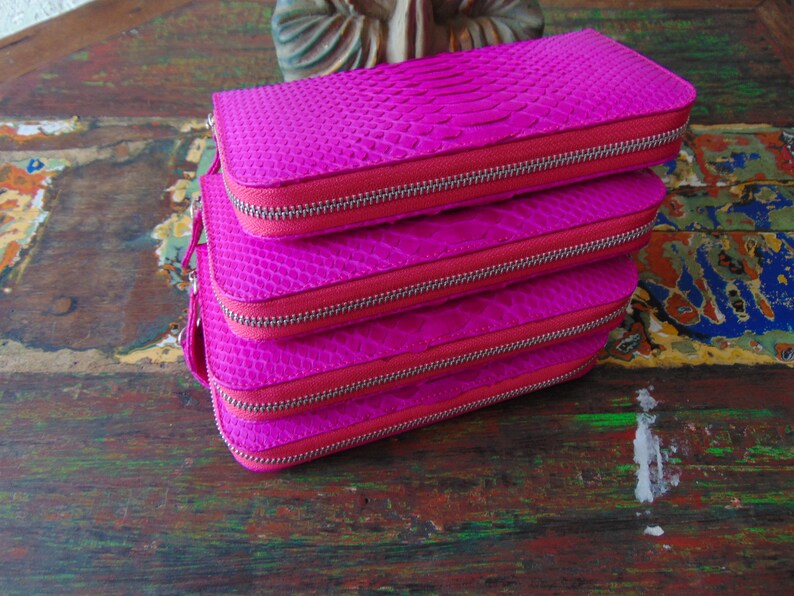 K&ouml;nnte beinhalten: Stapel von vier rosa Ledergeldb&ouml;rsen mit Rei&szlig;verschluss. Die Geldb&ouml;rsen haben ein strukturiertes Schlangenhautmuster.